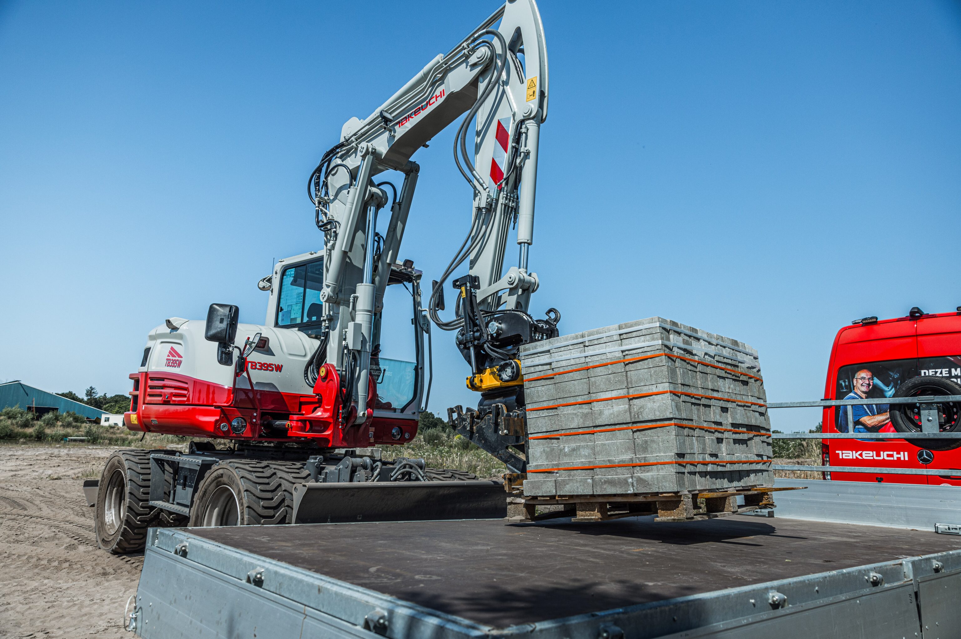 Een Takeuchi tb395w graafmachine met een palletvork aanbouwstuk tilt een pallet stenen van een aanhangwagen