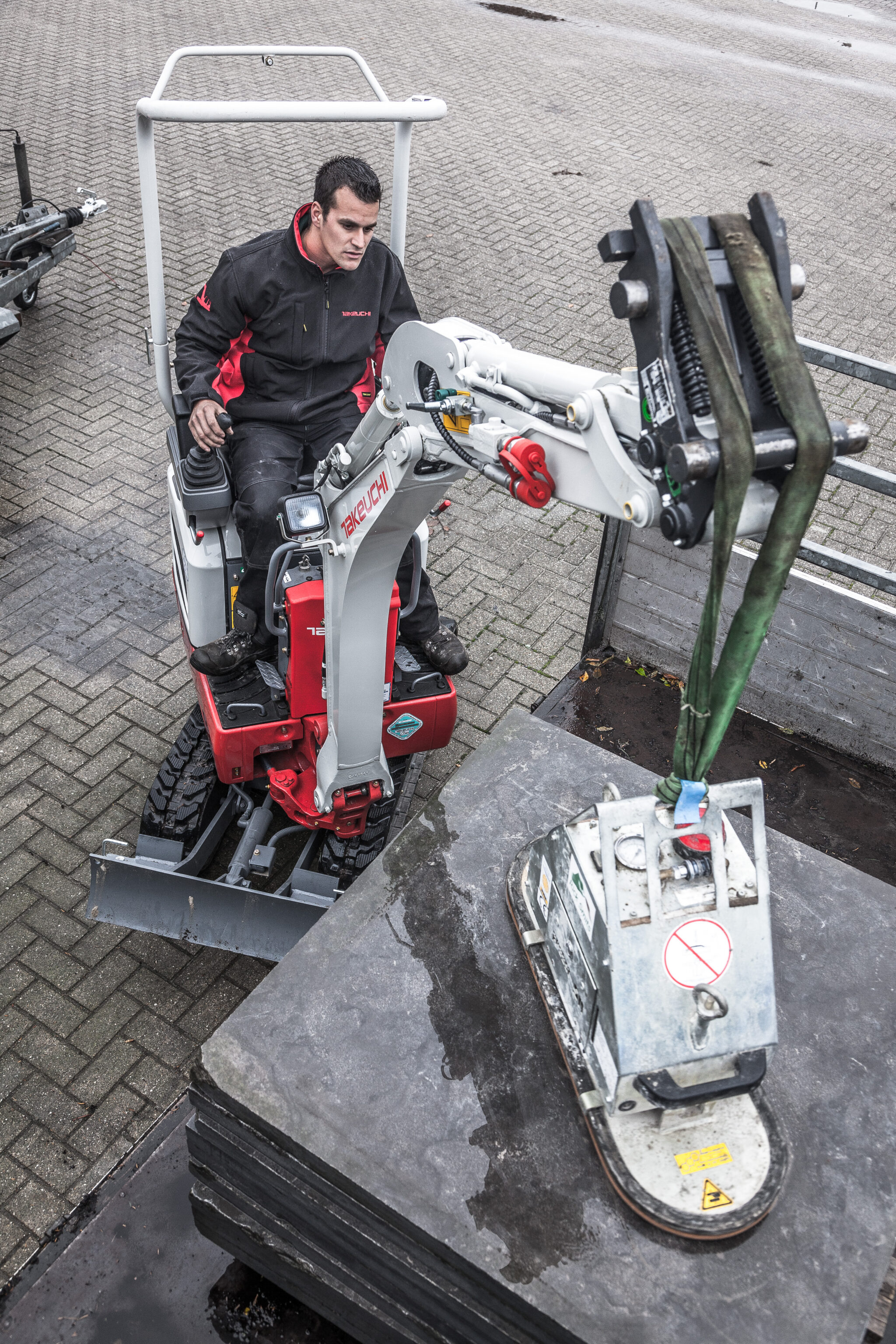 kleine Takeuchi graafmachin die met een vacuümunit een zware vierkante tegel verplaatst